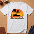 A white short-sleeve t-shirt laid flat on a cork background, featuring a retro-style graphic of two palm trees in front of an orange and yellow sunset with the text 'Dreaming of Summer Vacation NO MORE CLASSES'.