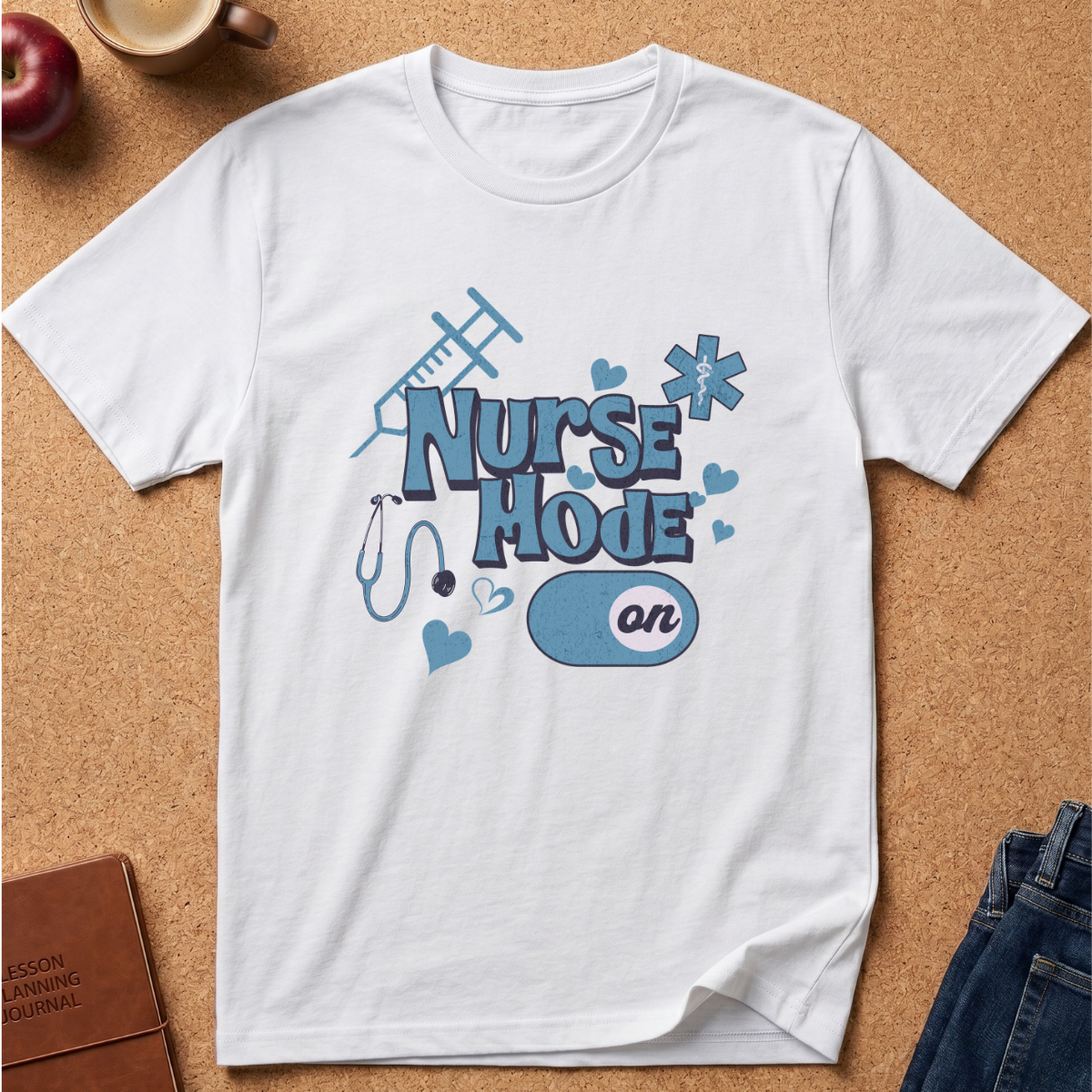 A white short-sleeve crewneck t-shirt with a 'Nurse Mode On' graphic in shades of blue, laid flat on a cork board background with lifestyle props like an apple, a coffee mug, and jeans.