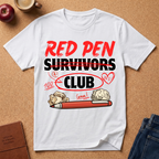 White t-shirt with 'Red Pen Survivors Club' design on a brown surface with accessories.