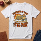 White t-shirt with a graphic design of a school bus on fire and text, placed on a brown surface with an apple and coffee cup. Teaching is easy It's like riding a bike.. except the Bike is on fire.