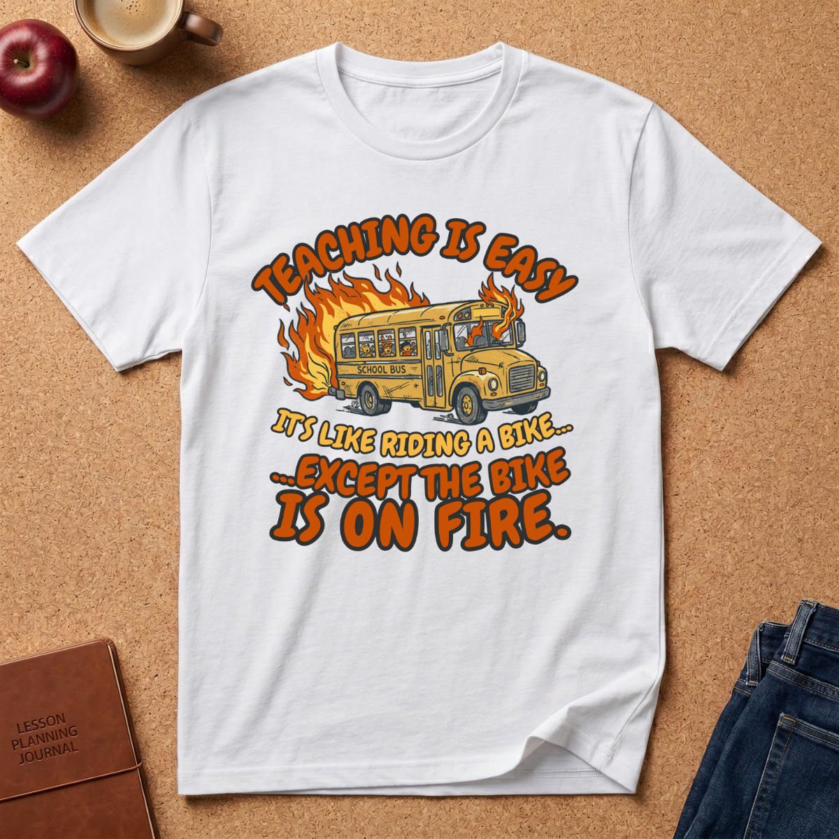 White t-shirt with a graphic design of a school bus on fire and text, placed on a brown surface with an apple and coffee cup. Teaching is easy It's like riding a bike.. except the Bike is on fire.