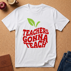 A white short-sleeve graphic T-shirt laid flat on a cork surface, featuring a green apple-leaf graphic above the red text 'TEACHERS GONNA TEACH.' Surrounding props include an apple, a coffee cup, a journal, and jeans.
