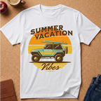A white short-sleeve crewneck t-shirt laid flat on a cork background, featuring a retro graphic of a green jeep with a surfboard on its roof, surrounded by a yellow-orange sun and the words 'SUMMER VACATION' and 'Vibes'.
