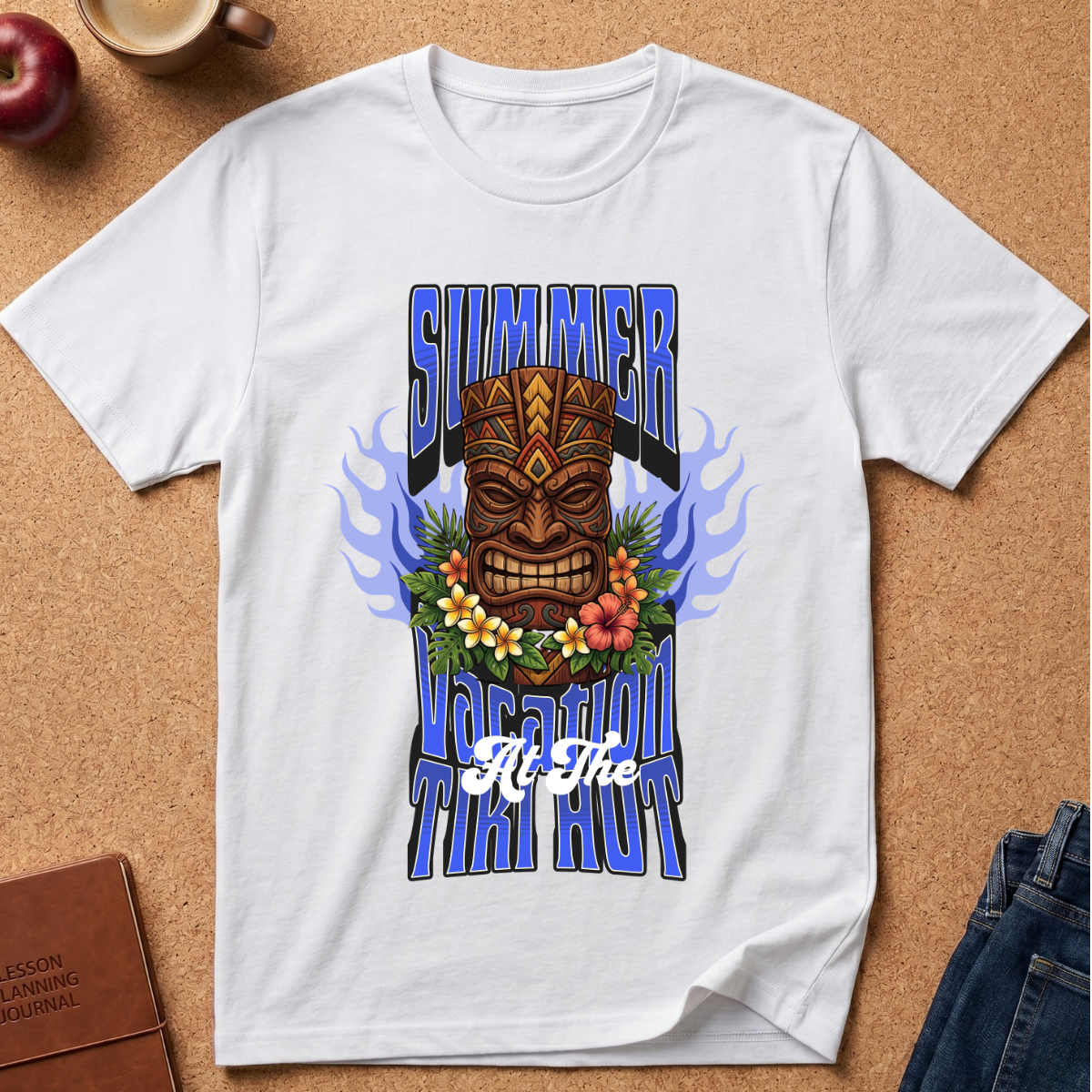 A white short-sleeve graphic T-shirt laid flat on a cork background, featuring a large tiki mask design with blue flame-like side elements, flowers across the chest, and stylized text reading 'SUMMER TUITI HOUR' plus 'All The'.