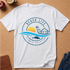 A white short-sleeve crewneck t-shirt laid flat on a cork board background, featuring a blue and yellow circular graphic that reads 'BEACH LIFE SUMMER VACATION' with a sunset, palm tree, beach house, waves, and fish.