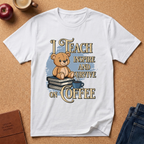 A white short-sleeve graphic T-shirt laid flat on a cork background, featuring a teddy bear sitting on books with the text 'I TEACH INSPIRE AND SURVIVE ON COFFEE.' Decorative props including an apple, a coffee cup, a journal, and jeans are partially visible around the shirt.